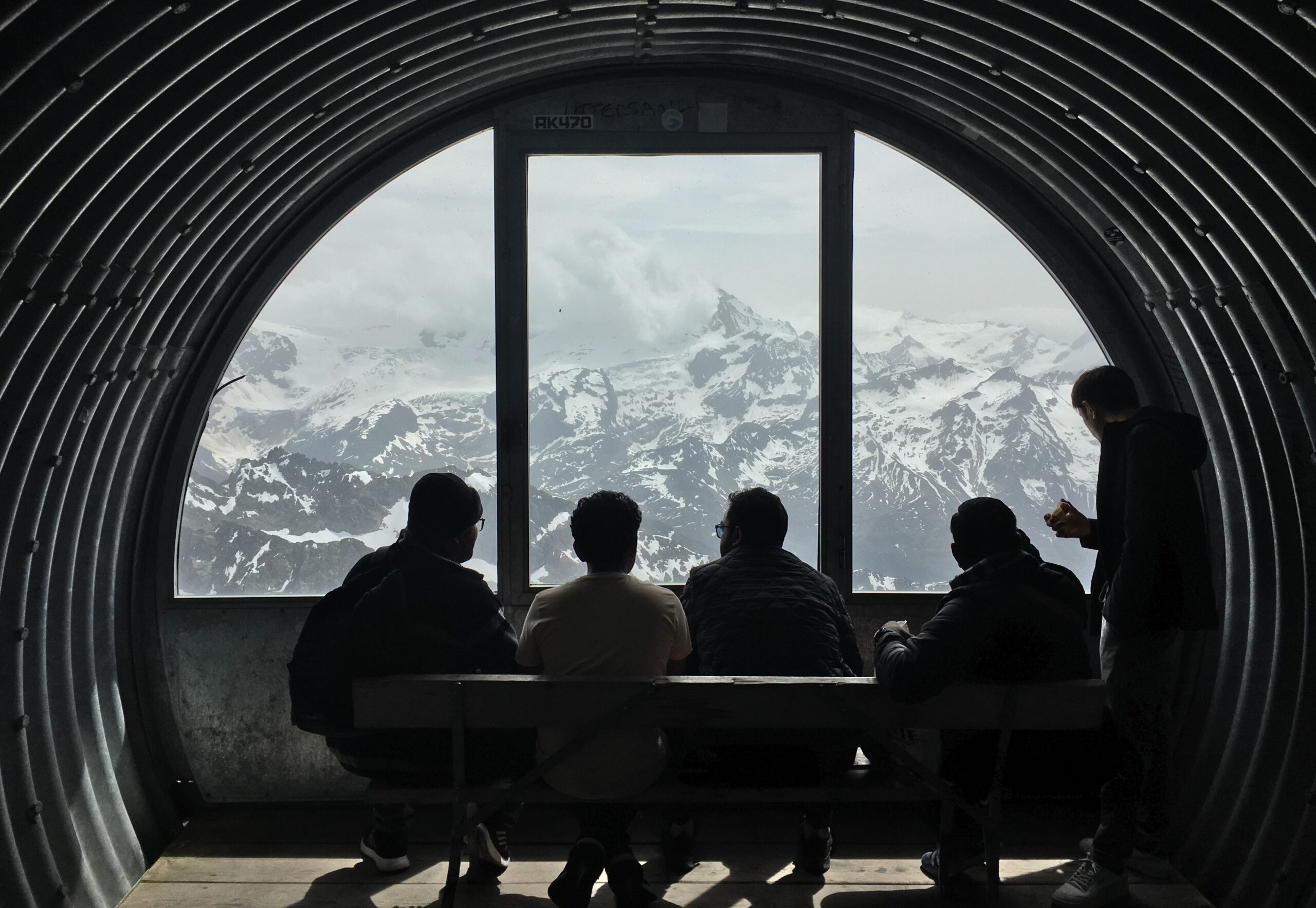A group of people sit on a bench in front of a circular window overlooking the Swiss Alps